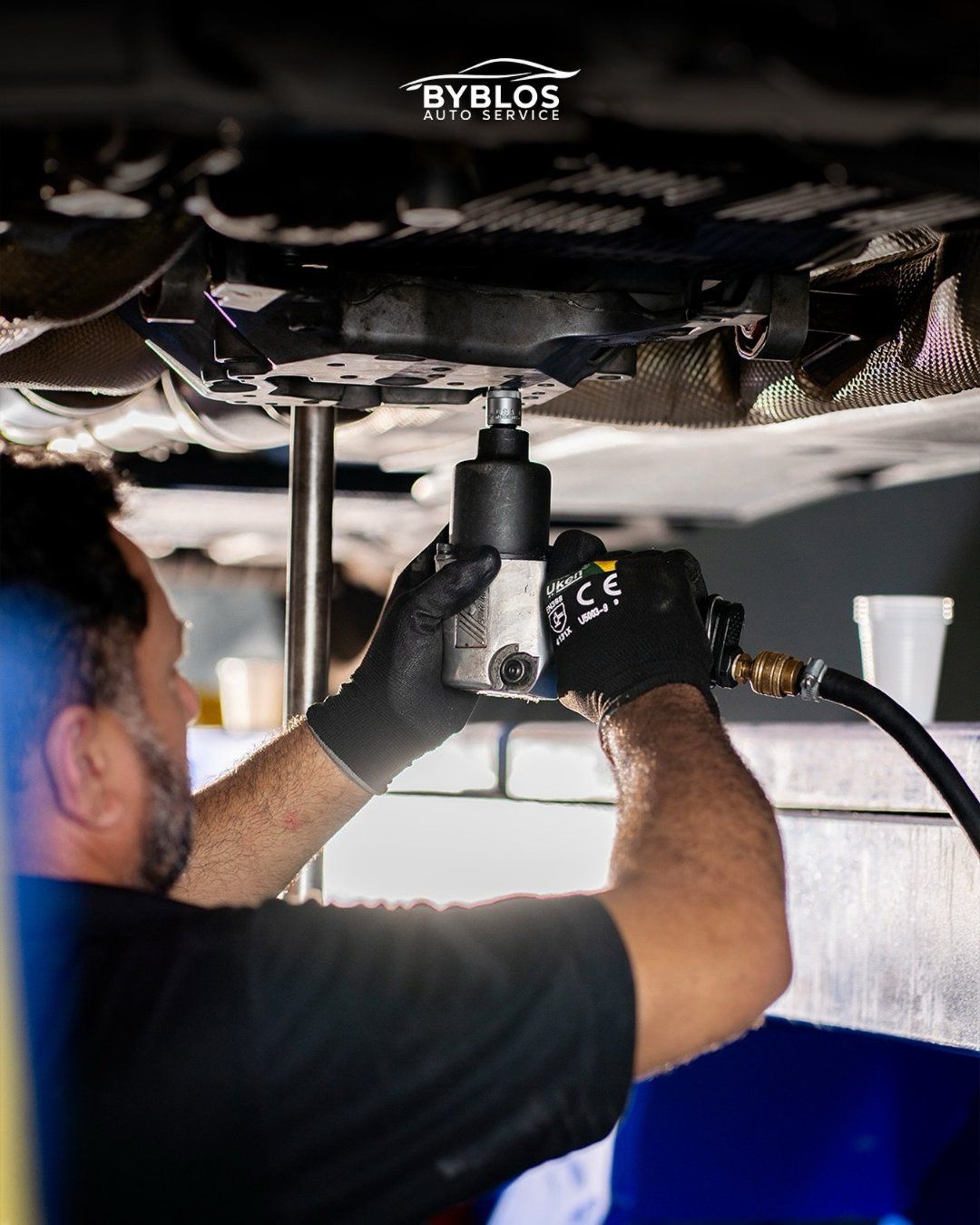 A technician in black gloves working under a raised car with an impact wrench